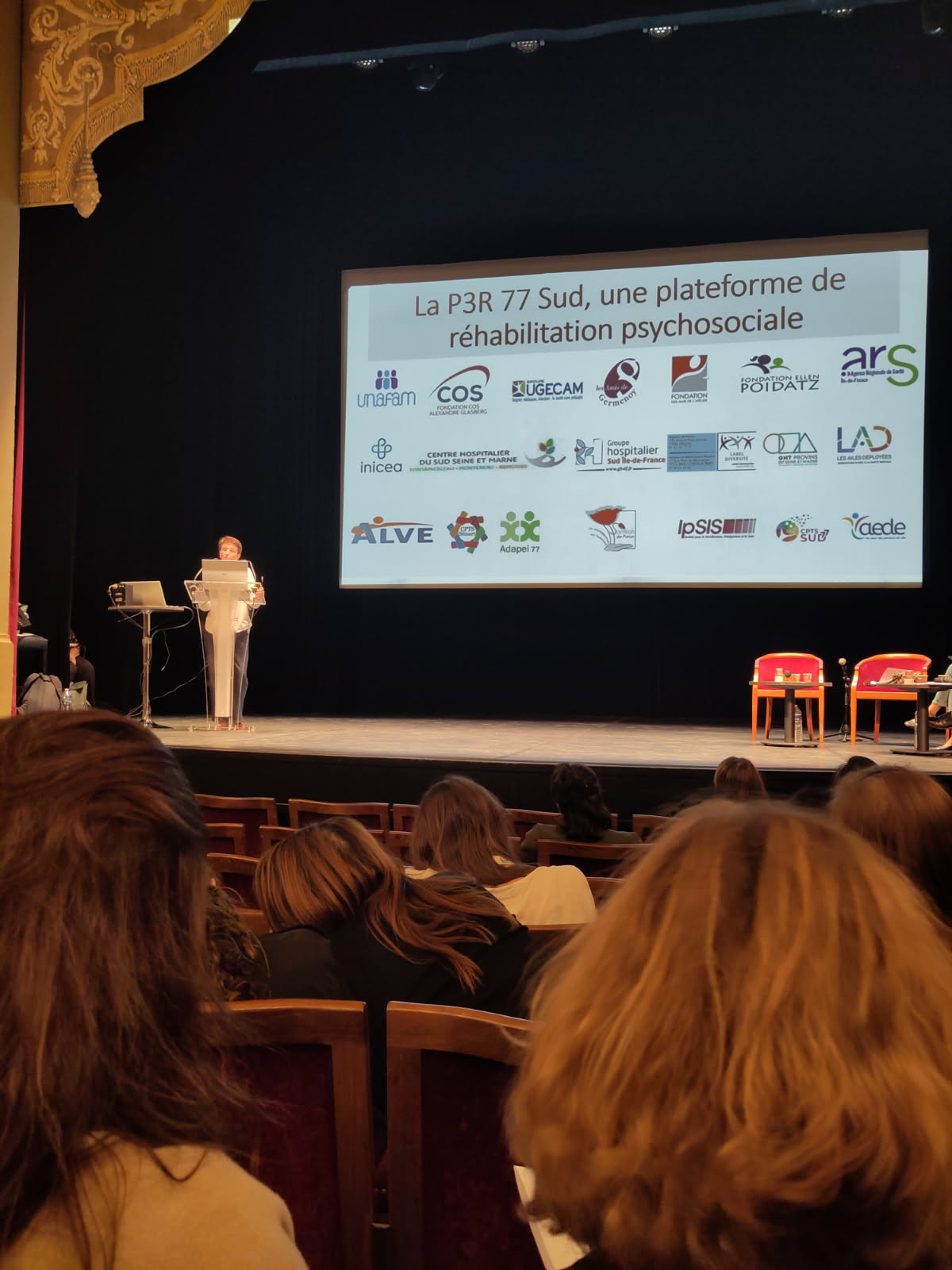 CRPF Conférence réhabilitation psychosociale Conférence dans un théâtre : une femme présente sur scène devant un public, avec une diapositive affichant des logos et le titre d’une plateforme de réhabilitation psychosociale.