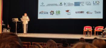Conférence dans un théâtre : une femme présente sur scène devant un public, avec une diapositive affichant des logos et le titre d’une plateforme de réhabilitation psychosociale.