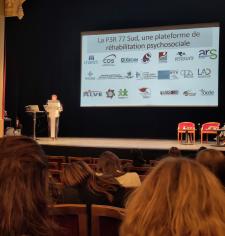 CRPF Conférence réhabilitation psychosociale Conférence dans un théâtre : une femme présente sur scène devant un public, avec une diapositive affichant des logos et le titre d’une plateforme de réhabilitation psychosociale.